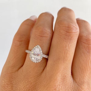 May include: A pear-shaped engagement ring with a halo setting. The ring is made of silver and has a clear center stone.
