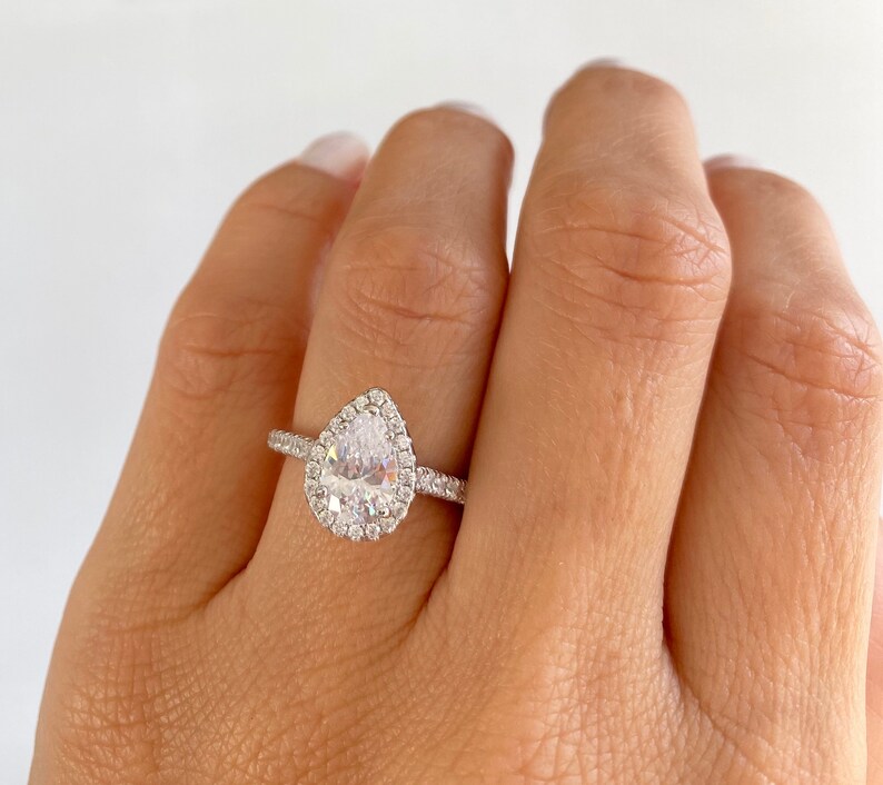 May include: A pear-shaped engagement ring with a halo setting. The ring is made of silver and has a clear center stone.
