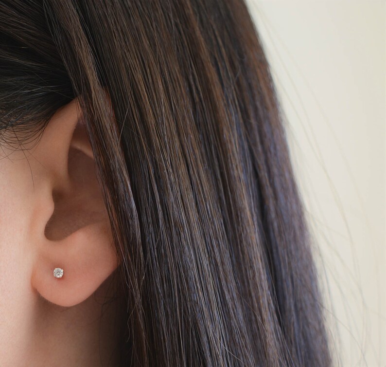 May include: A close-up of a person's ear with a small, round, clear diamond stud earring.