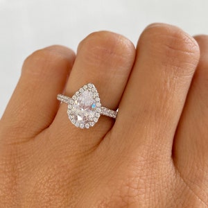 May include: A pear-shaped engagement ring with a halo setting. The ring is set in silver and features a large, clear center stone surrounded by smaller diamonds.