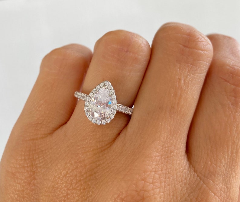 May include: A pear-shaped engagement ring with a halo setting. The ring is set in silver and features a large, clear center stone surrounded by smaller diamonds.