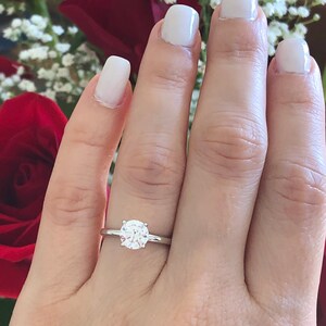 May include: A close-up of a hand wearing a silver ring with a round, clear diamond solitaire.