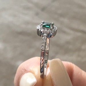 May include: A silver ring with a green emerald gemstone surrounded by smaller clear gemstones. The ring has a delicate band with small clear gemstones.