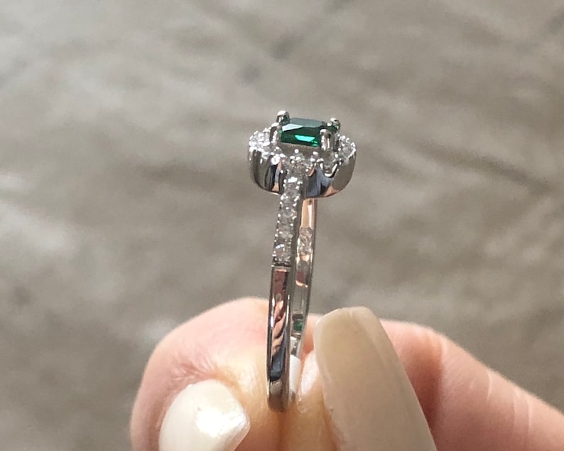May include: A silver ring with a green emerald gemstone surrounded by smaller clear gemstones. The ring has a delicate band with small clear gemstones.
