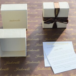 May include: Three white gift boxes with the word "Jadorelli" printed on them. The boxes are on brown paper with the same word printed repeatedly. A white card with the text "With love from Jadorelli" and "Care Tips" is also on the paper.