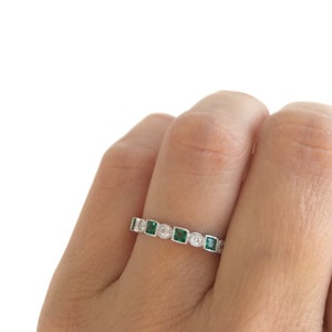 May include: A silver ring featuring a series of square emerald-green gemstones and round clear gemstones. The ring is set with alternating emeralds and clear stones, creating a sparkling band. The ring is shown on a finger.