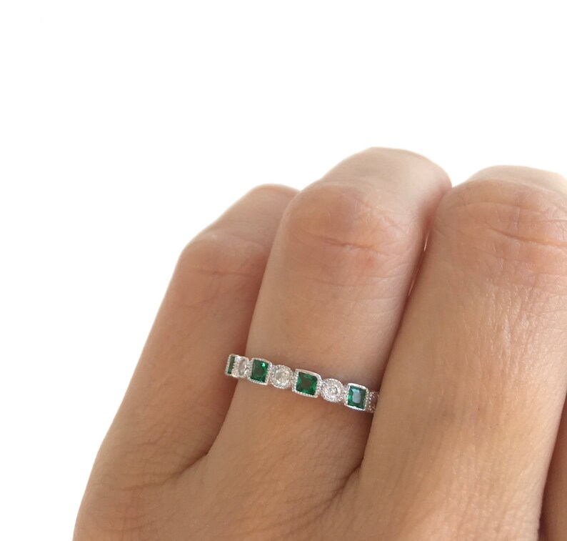 May include: A silver ring featuring a series of square emerald-green gemstones and round clear gemstones. The ring is set with alternating emeralds and clear stones, creating a sparkling band. The ring is shown on a finger.