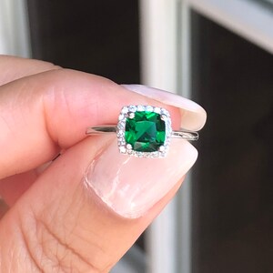 Puede incluir: Un anillo de plata con una piedra preciosa cuadrada de color verde esmeralda rodeada por un halo de piedras preciosas m&aacute;s peque&ntilde;as y transparentes.