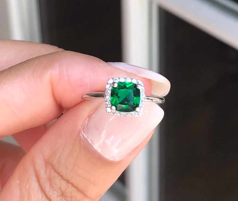 Puede incluir: Un anillo de plata con una piedra preciosa cuadrada de color verde esmeralda rodeada por un halo de piedras preciosas m&aacute;s peque&ntilde;as y transparentes.