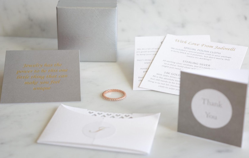 May include: Rose gold eternity ring with small clear stones, displayed with a gift box, cards, and an envelope. The cards include text in gold lettering. The envelope has a heart design and a monogram. The ring is a delicate piece of jewelry.