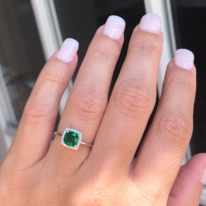 Puede incluir: Un anillo de plata con una piedra preciosa cuadrada verde esmeralda rodeada por un halo de piedras preciosas m&aacute;s peque&ntilde;as y transparentes.