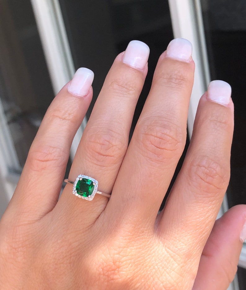 Puede incluir: Un anillo de plata con una piedra preciosa cuadrada verde esmeralda rodeada por un halo de piedras preciosas m&aacute;s peque&ntilde;as y transparentes.