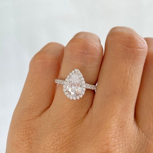May include: A pear-shaped engagement ring with a halo setting. The ring is set in silver and features a large, clear center stone surrounded by smaller, clear stones.