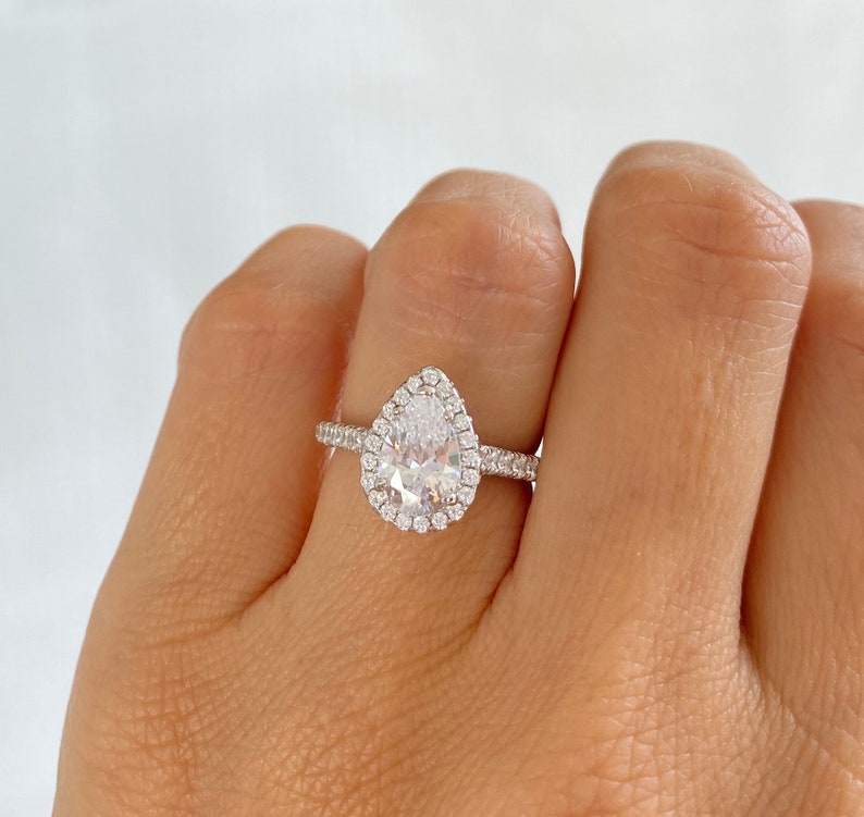 May include: A pear-shaped engagement ring with a halo setting. The ring is set in silver and features a large, clear center stone surrounded by smaller, clear stones.