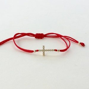 May include: A red string bracelet with a gold cross charm adorned with clear cubic zirconia stones.