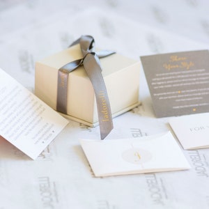 May include: A small, square, cream-colored gift box with a gray satin ribbon tied in a bow. The box is labeled "Jador&eacute;lli" in gold lettering. The box is sitting on a white surface with several small cards, one of which says "Special Occasions" and another says "For You".