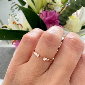 Op de afbeelding: Een ring van roségoud met twee kleine diamanten op de band. De ring is aan één kant open en wordt aan een vinger gedragen.