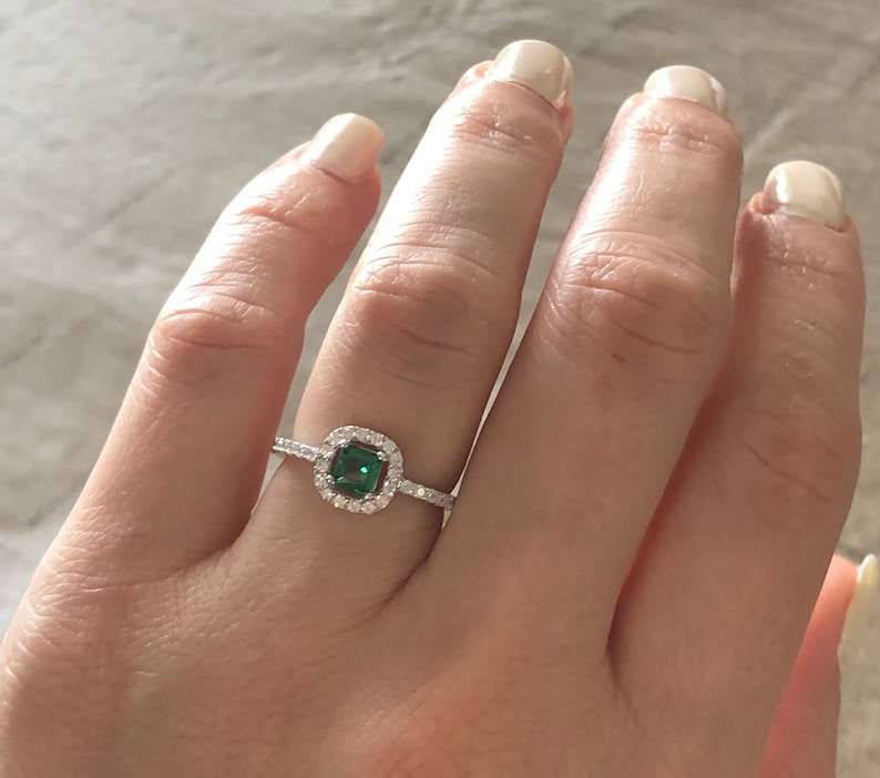 May include: A silver ring with a square emerald green gemstone surrounded by smaller clear gemstones.