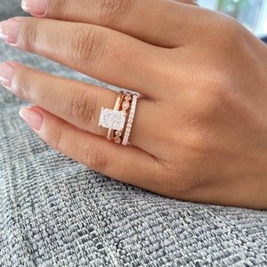 Puede incluir: Un anillo de compromiso de oro rosa con una piedra central rectangular grande y dos bandas de diamantes más pequeñas.