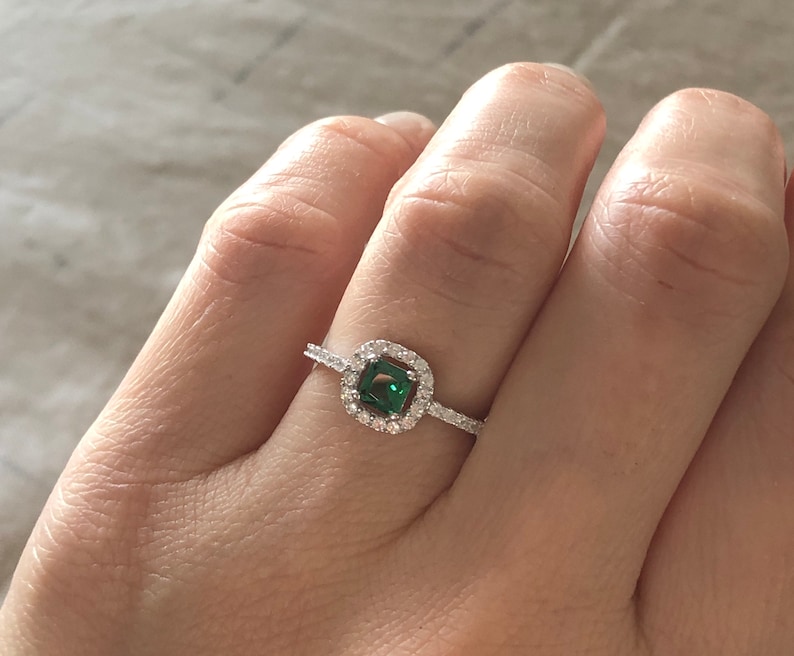 May include: A silver ring with a square emerald green gemstone surrounded by smaller clear gemstones.