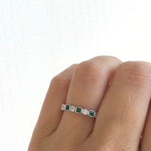 May include: A silver ring featuring a series of alternating emerald-cut green gemstones and round white gemstones. The ring is set with small, round accent stones along the band, creating a delicate and elegant design.