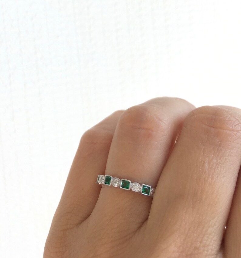 May include: A silver ring featuring a series of alternating emerald-cut green gemstones and round white gemstones. The ring is set with small, round accent stones along the band, creating a delicate and elegant design.