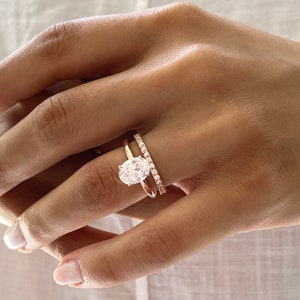 May include: A close-up of a hand wearing two rings. The top ring features a large oval-shaped diamond, while the bottom ring is a thin band with small diamonds set in a pave setting. Both rings are made of rose gold.