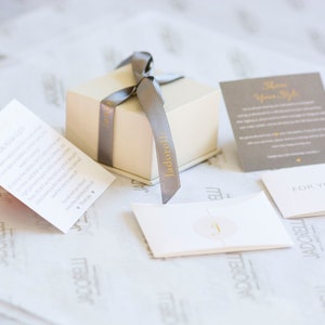 May include: A small, cream-colored gift box with a gray ribbon tied around it. The box is labeled "Jadore It" in gold lettering. The box is sitting on a white, textured surface with several small cards. One card says "Show Your Style" and another says "For You".