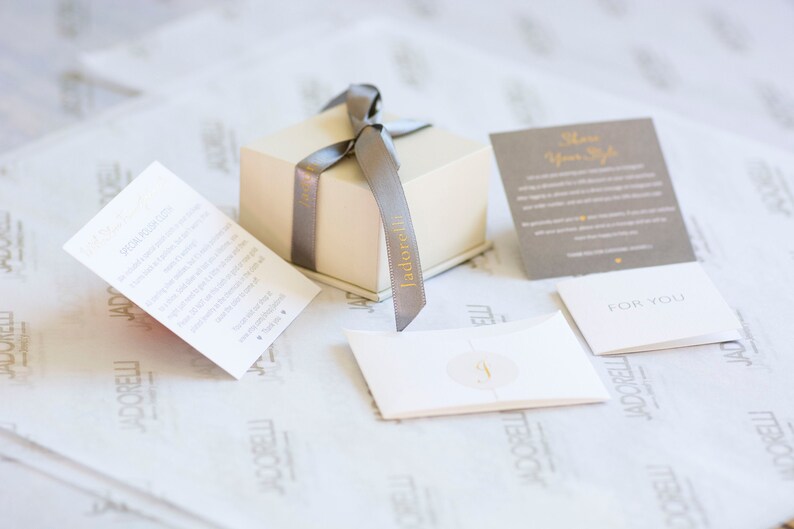 May include: A small, cream-colored gift box with a gray ribbon tied around it. The box is labeled "Jadore It" in gold lettering. The box is sitting on a white, textured surface with several small cards. One card says "Show Your Style" and another says "For You".