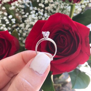 May include: A silver engagement ring with a large, clear diamond set in a prong setting. The ring is held by a hand against a backdrop of red roses.