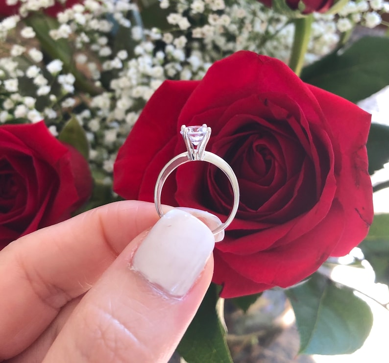 May include: A silver engagement ring with a large, clear diamond set in a prong setting. The ring is held by a hand against a backdrop of red roses.