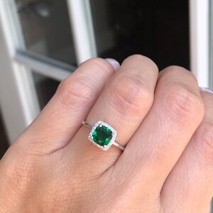 Puede incluir: Un anillo de plata con una piedra preciosa cuadrada de color verde esmeralda rodeada por un halo de piedras preciosas m&aacute;s peque&ntilde;as y transparentes.