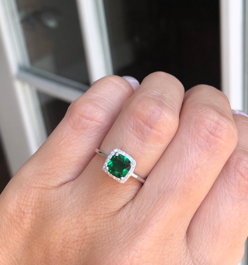 Puede incluir: Un anillo de plata con una piedra preciosa cuadrada de color verde esmeralda rodeada por un halo de piedras preciosas m&aacute;s peque&ntilde;as y transparentes.