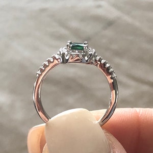 May include: A silver ring with a green emerald gemstone surrounded by smaller clear gemstones. The band is set with smaller clear gemstones.