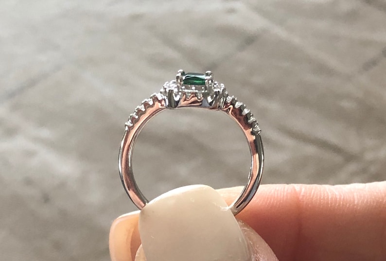 May include: A silver ring with a green emerald gemstone surrounded by smaller clear gemstones. The band is set with smaller clear gemstones.