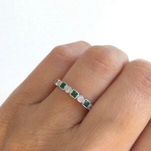 May include: A silver ring featuring alternating emerald-cut green gemstones and round, clear gemstones. The ring is set with small, clear accent stones along the band. The ring is shown on a person's finger.