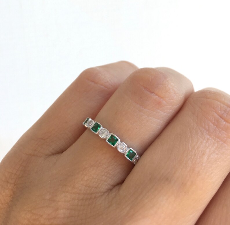 May include: A silver ring featuring alternating emerald-cut green gemstones and round, clear gemstones. The ring is set with small, clear accent stones along the band. The ring is shown on a person's finger.