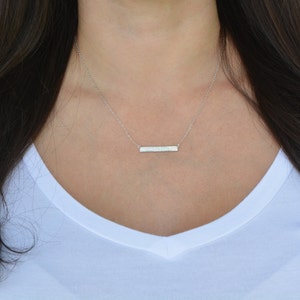 May include: A silver bar necklace worn by a model in a white top.