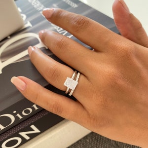 Peut inclure: Gros plan d'une main portant une bague en argent avec un grand diamant rectangulaire transparent. La bague est sertie de petits diamants autour de la pierre centrale. La bague est au troisième doigt de la main gauche.