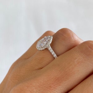 May include: A pear-shaped engagement ring with a halo setting and a pave band. The ring is set in silver and features a large, clear center stone.