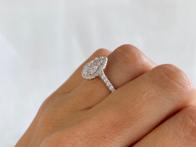 May include: A pear-shaped engagement ring with a halo setting and a pave band. The ring is set in silver and features a large, clear center stone.