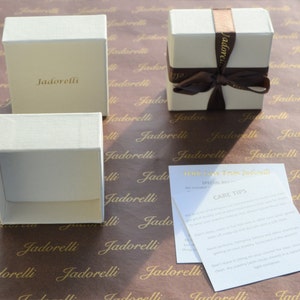 Op de afbeelding: Drie witte cadeaudozen met het woord "Jadorelli" erop gedrukt. De dozen liggen op bruin papier met hetzelfde woord herhaaldelijk erop gedrukt. E&eacute;n doos is vastgebonden met een bruin lint. Een witte kaart met de tekst "With love from Jadorelli" en "Special Offer" en "Care Tips" ligt op het papier.