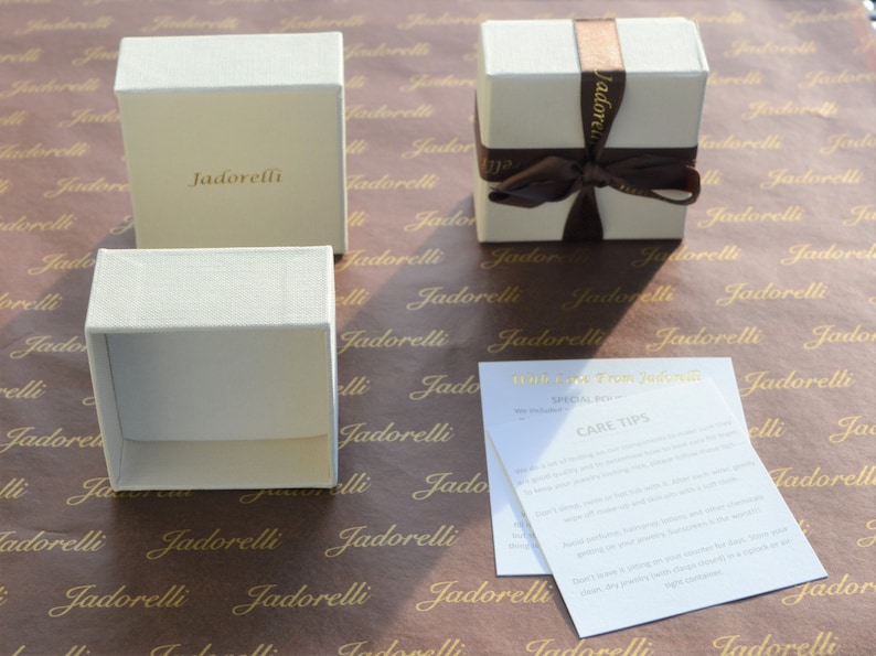 Op de afbeelding: Drie witte cadeaudozen met het woord "Jadorelli" erop gedrukt. De dozen liggen op bruin papier met hetzelfde woord herhaaldelijk erop gedrukt. E&eacute;n doos is vastgebonden met een bruin lint. Een witte kaart met de tekst "With love from Jadorelli" en "Special Offer" en "Care Tips" ligt op het papier.