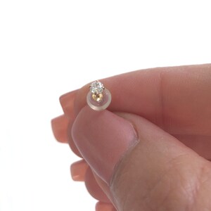 May include: A single clear diamond stud earring with a gold setting. The earring is held in a person's hand.