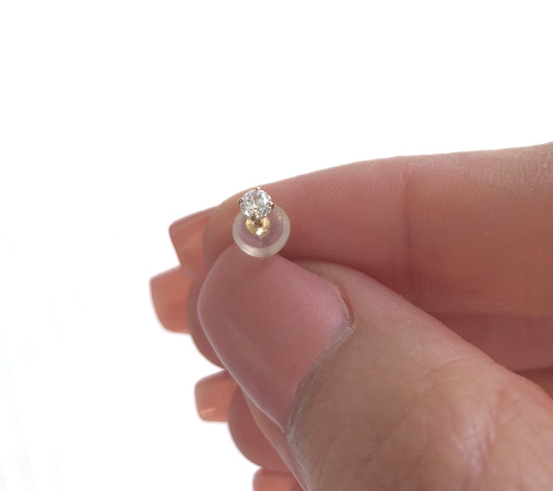 May include: A single clear diamond stud earring with a gold setting. The earring is held in a person's hand.