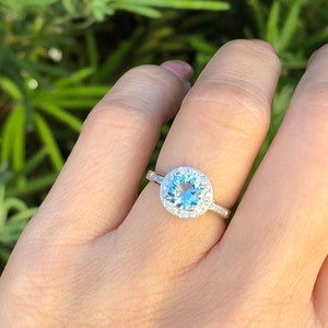 Pode incluir: Um anel de prata com uma gema de topázio azul redonda cercada por um halo de diamantes menores.
