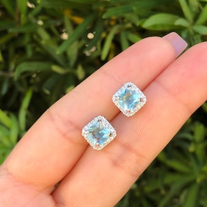 Pode incluir: Um par de brincos de prata com pedras azuis quadradas rodeadas por um halo de pedras transparentes menores.