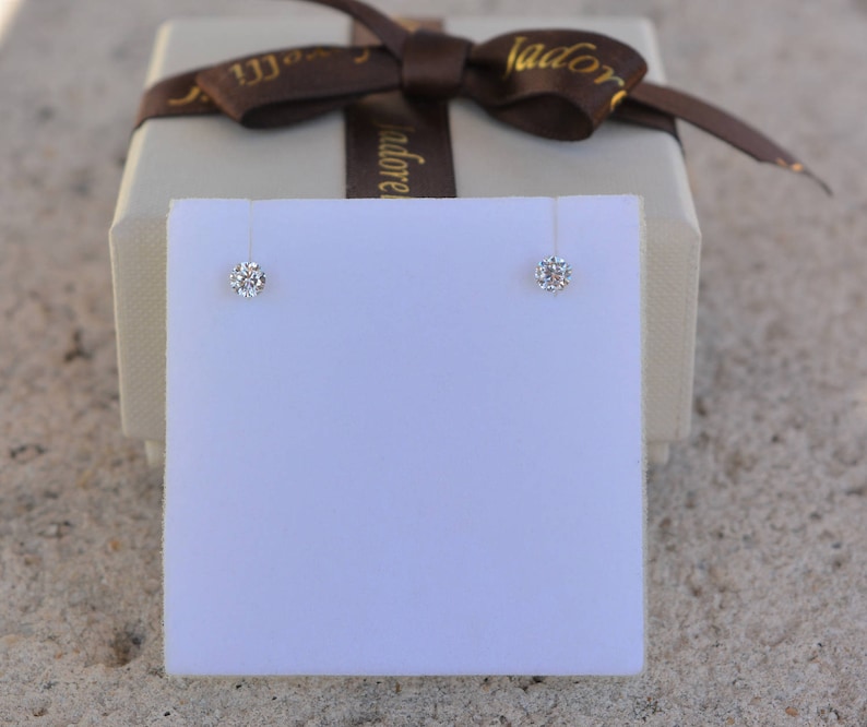 May include: A pair of diamond stud earrings displayed on a white card, with a brown gift box in the background.