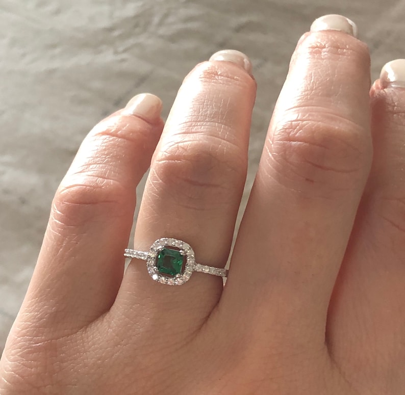 May include: A silver ring with a square emerald green gemstone surrounded by a halo of smaller clear gemstones.