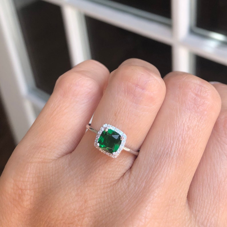 Puede incluir: Un anillo de plata con una piedra preciosa cuadrada verde esmeralda rodeada por un halo de piedras preciosas m&aacute;s peque&ntilde;as y transparentes.